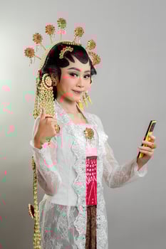 An Indonesian bride in Solo Putri attire smiles confidently while holding a smartphone and giving a thumbs-up. She wears an ornate golden headdress, elegant lace kebaya, and intricate jewelry. photo