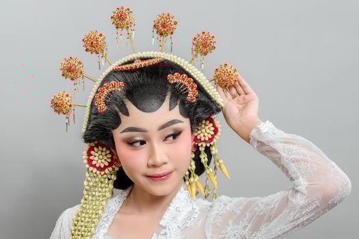 A Indonesian bride in Solo Putri attire poses elegantly, lifting her hand with a confident smile. She wears an intricate golden headdress, jasmine garland, and white lace kebaya.. photo