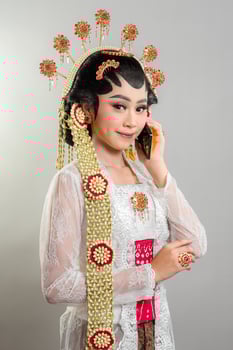 An Indonesian bride in Solo Putri attire gracefully talks on a smartphone. She wears an elegant golden headdress, intricate jewelry, a white lace kebaya, and traditional Javanese accessories. photo