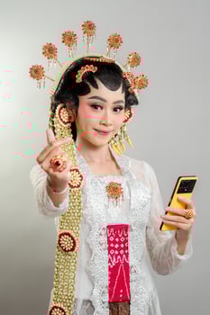 A Indonesian bride in Solo Putri attire smiles while holding a smartphone. She wears an elegant golden headdress, intricate jewelry, a white lace kebaya, and traditional Javanese accessories. photo