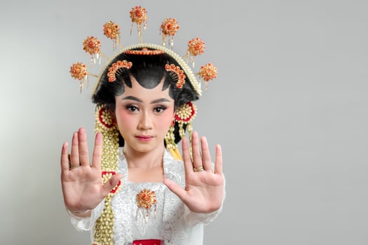 An Indonesian bride in Solo Putri attire, wearing an ornate headdress and lace kebaya. She raises both hands in a stop gesture with a serious expression, exuding elegance and grace. photo