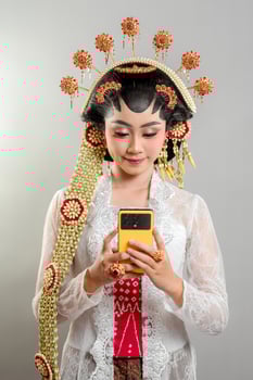 An Indonesian bride in Solo Putri attire elegantly interacts with her smartphone. Wearing a golden headdress jasmine accessories, and a lace kebaya, she smiles while looking focused. photo