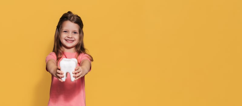 A smiling girl with healthy teeth holds a plastic tooth in her hands on a yellow insulated background. A place for your text. Oral hygiene. Pediatric dentistry. Prosthetics. Rules for brushing teeth,. photo