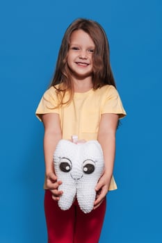 A smiling girl with healthy teeth holds a tooth toy in her hands on a blue isolated background. Oral hygiene. Pediatric dentistry. Prosthetics. Rules for brushing teeth. Vertical photo. photo