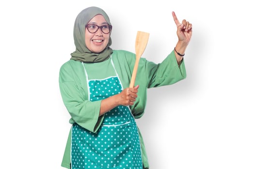 Cheerful Muslim woman wearing an apron, smiling while holding a wooden spatula, representing home cooking, culinary passion, and a joyful kitchen atmosphere photo