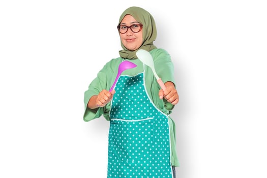 Cheerful Muslim woman in apron holding kitchen utensils, ready to cook meals photo