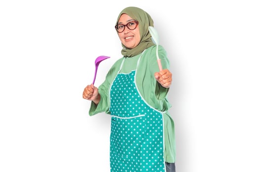 Happy Muslim woman in apron holding kitchen utensils, ready to cook delicious meals photo