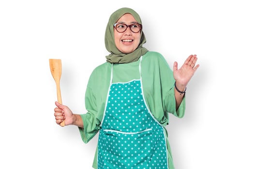 Cheerful Muslim woman chef wearing an apron, holding a wooden spatula, looking excited to cook in a home kitchen setting photo