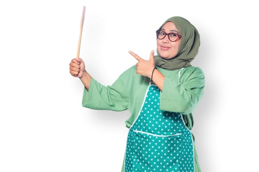 Cheerful Muslim woman in a hijab and apron holding a wooden spoon and pointing with confidence photo