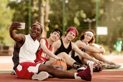 Team of happy basketball players taking selfie while relaxing after game at outdoor stadium, blank space. Group of multiracial sportsmen making photo of themselves on break