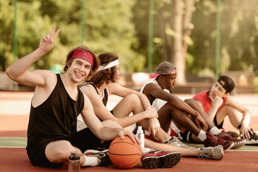 Happy young basketball player and his multinational team having rest at outdoor arena, copy space. Cheerful millennial guy showing peace gesture at camera, relaxing with his fellow sportsmen outside photo