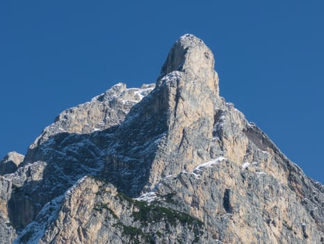 A rugged mountain peak with sharp rock formations and snow patches, set against a clear blue sky. Greenery accents the lower slopes in an alpine setting. photo