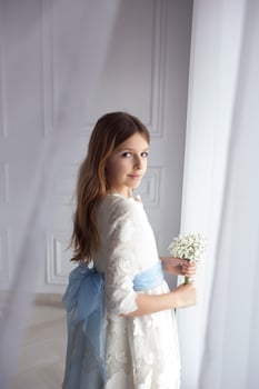 beautiful girl in elegant white dress with bouquet of flowers, looking out the window during first communion photo
