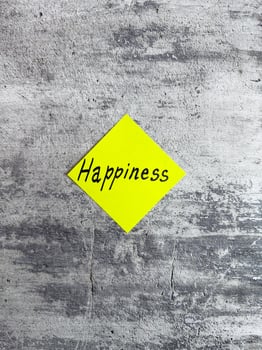 Vertical photograph showcasing a bright yellow diamond-shaped sticky note with the word Happiness against a textured grey background, symbolizing joy and positivity in daily life photo