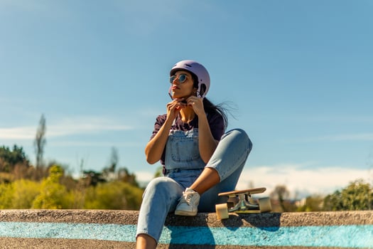 Latin skater girl fastening her helmet sitting on a wall with her surfskate next to her photo