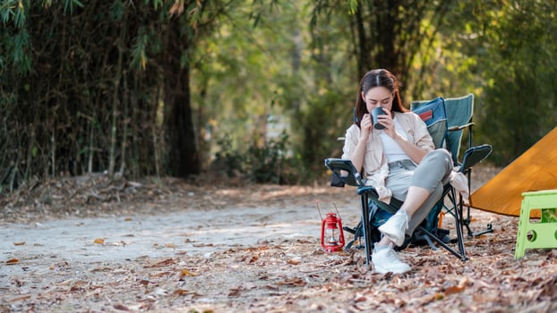 A woman relaxes with a coffee in a forest campsite, surrounded by autumn leaves and camping equipment. photo