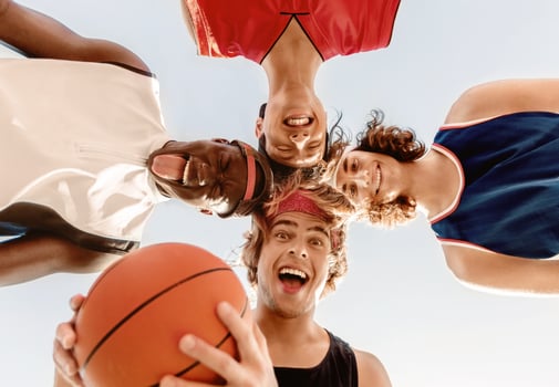 Diverse basketball players putting their heads together and making silly faces, having fun before game, outdoors. Bottom view photo