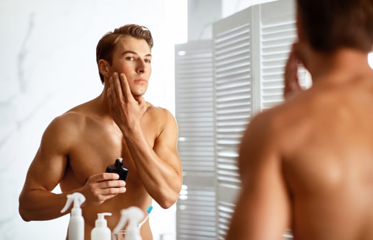 Charming man applying lotion on his face after shaving, looking in mirror, standing at the bath room photo