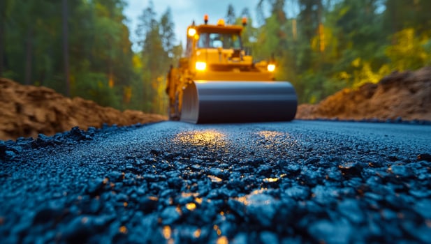 Machines lay asphalt at dawn. A road roller compacting freshly laid asphalt in a forested area during early morning light, creating a smooth surface. photo