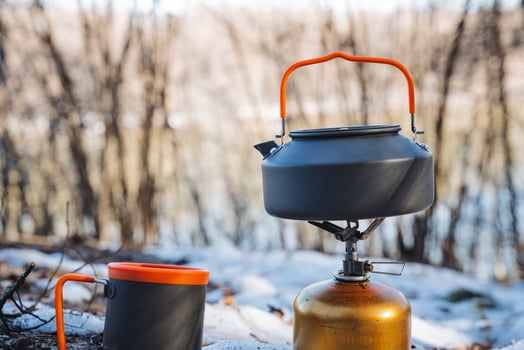 A kettle made of metal and a ceramic cup are resting on top of a gas stove situated in a snowy outdoor setting, surrounded by a blanket of white snowflakes in the chilly air photo
