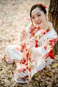 Cheerful Asian woman in traditional floral kimono holding Japanese dango skewers toward camera with smile, enjoying sweet treat outdoors in spring season photo