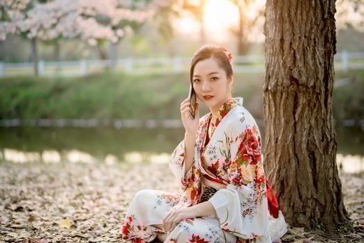 Elegant Asian woman in traditional floral kimono talking on smartphone under tree during sunset, blending Japanese heritage with modern digital lifestyle photo