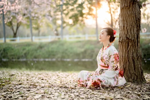 Asian woman in traditional floral kimono meditating under tree by the river during soft sunrise light, peaceful nature and harmony in Japanese culture scene photo