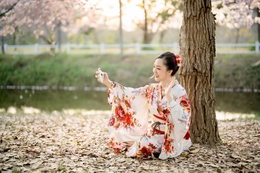 Asian woman in traditional floral kimono calling with smartphone under cherry blossom tree in natural setting, blending tradition and modern lifestyle photo