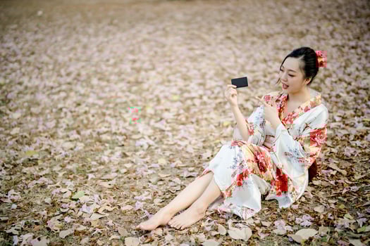 Cheerful Asian woman in floral kimono sitting on dry leaf ground, pointing at black card, traditional Japanese outfit and natural outdoor setting photo