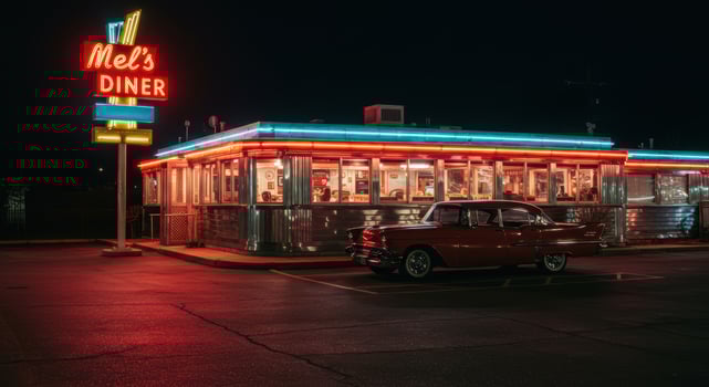 A vintage red car sits outside of a brightly lit retro diner at night. photo