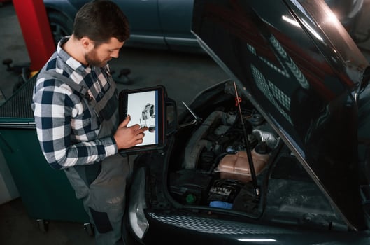 Standing ,,near the car with hood opened. Holding tablet. Man in uniform is working in the auto salon photo