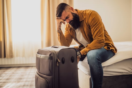 Man in his 30s sitting on bed in hotel room. He is tired and exhausted. Suitcase is beside the bed. photo