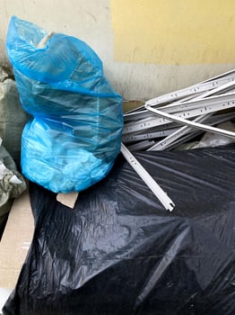 In urban alley filled with discarded materials, blue plastic bag atop large black bag. Surrounding debris includes sheets of metal and cardboard contrasting with faded yellow wall. Environment saving photo