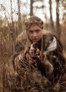 close-up of blonde girl in historical warrior costume, she aims with pistol. antique pistol in her hands. dressed in long brown fur coat, vintage dress. photo