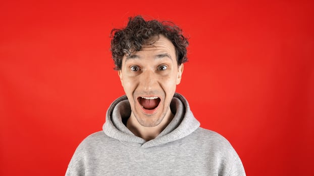 large close up portrait young curly man, joyfully surprised, holding his face with mouth open, pronouncing word wow on studio red background. Concept - discounts, Black Friday, excitement, advertising photo