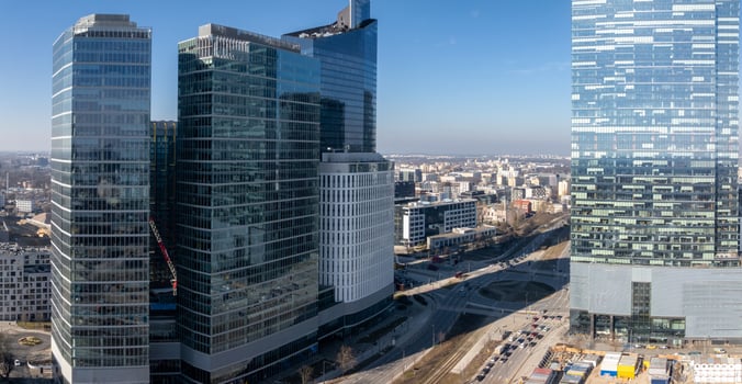 Modern skyscrapers with glass facades in Warsaw, Poland, surrounded by roads, smaller buildings, and ongoing construction under a clear blue sky. photo