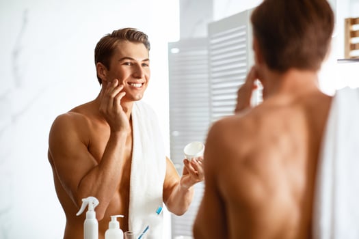 Male Facial Skincare Concept. Smiling Handsome Man Applying Cream On Smooth Face Caring For Skin After Shaving Routine, Looking In The Mirror, Standing Shirtless In Bathroom At Home photo