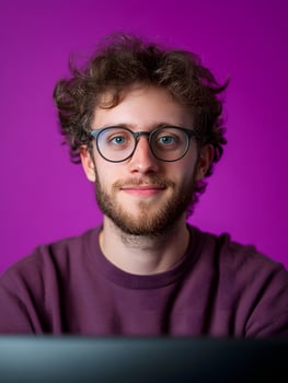 Portrait of a young smiling man with curly hair and glasses dressed casually and looking directly at the against a vibrant purple background suggesting his career as a game writer or developer photo