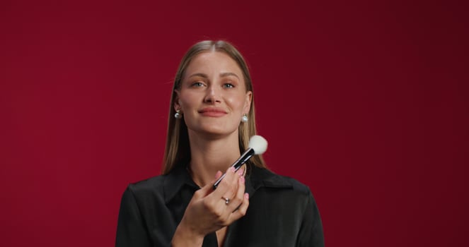 A woman in formal clothes applies blush to her face with a brush. She smiles at the camera photo