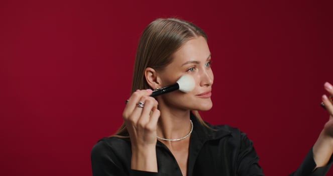 A woman in formal clothes applies blush to her face with a brush. She smiles at the camera while standing in the studio photo