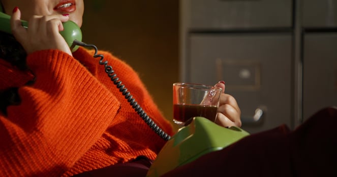A woman is talking on a wired phone and drinking coffee while sitting in a retro office photo