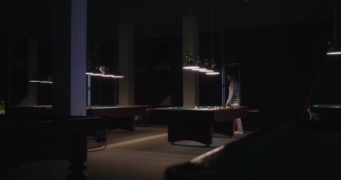 A young man of European appearance with a beard, dressed in casual clothes, plays billiards alone under dim lights in a billiard club - European photo