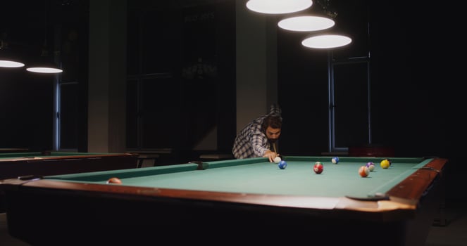 A young man of European appearance with a beard, dressed in casual clothes, plays billiards alone under dim lights in a billiard club - lights photo