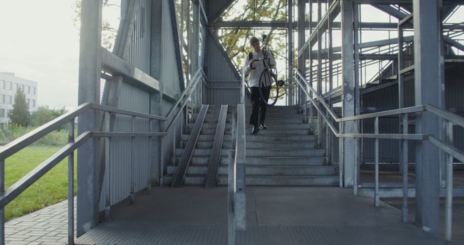 A young man of European appearance in a knitted hat with a backpack on his back rides a bicycle to a metal ladder, dismounts, takes a bicycle in his arms and carries it up the steps, bicycle photo
