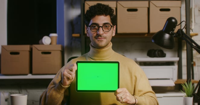 A young man looks directly into the camera and shows a tablet with green screen photo