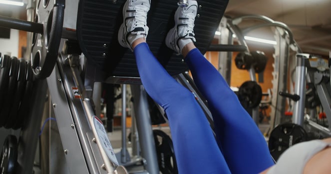 Blonde woman, athletic physique, dressed in sportswear, slowly lifts weights with her legs on the exercise machine, working out in the gym alone. Woman's face is blurred, focus on woman's legs photo