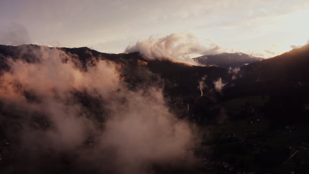 Panoramic view of the cottage town, located in a lowland between the Alpine mountains, the tops of which are covered with white clouds. Alps, Austria, drone photo