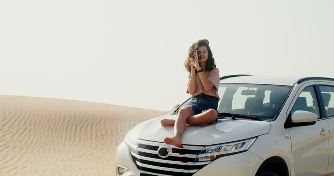 A woman takes a photo by sitting on the hood of a car in the desert