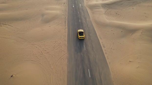 The drone flies over a yellow convertible driving along the road past the sand dunes of the desert - desert photo