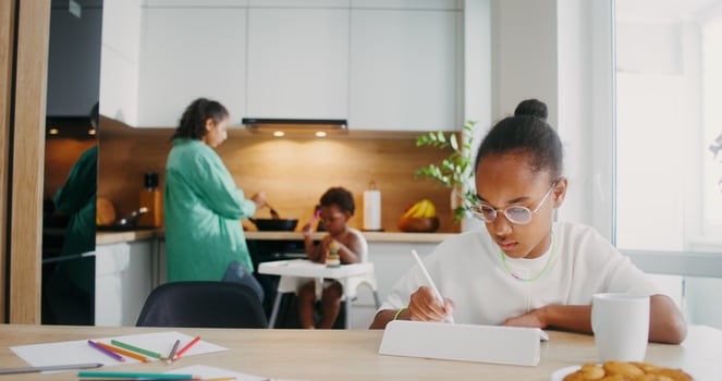 A girl draws with a stylus on a tablet while sitting at the kitchen table at home. Her family in the background is out of focus - family photo